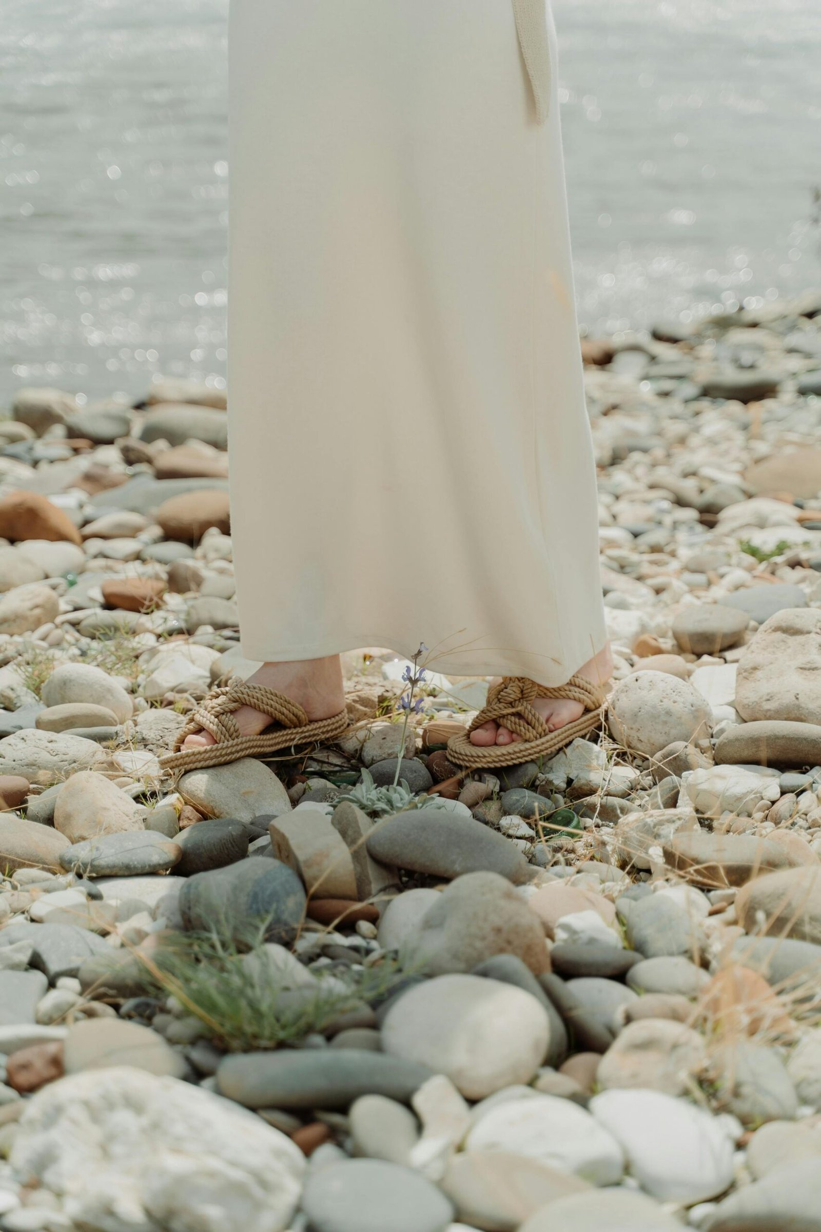 Person wearing natural rope sandals standing on a rocky shoreline in a light dress